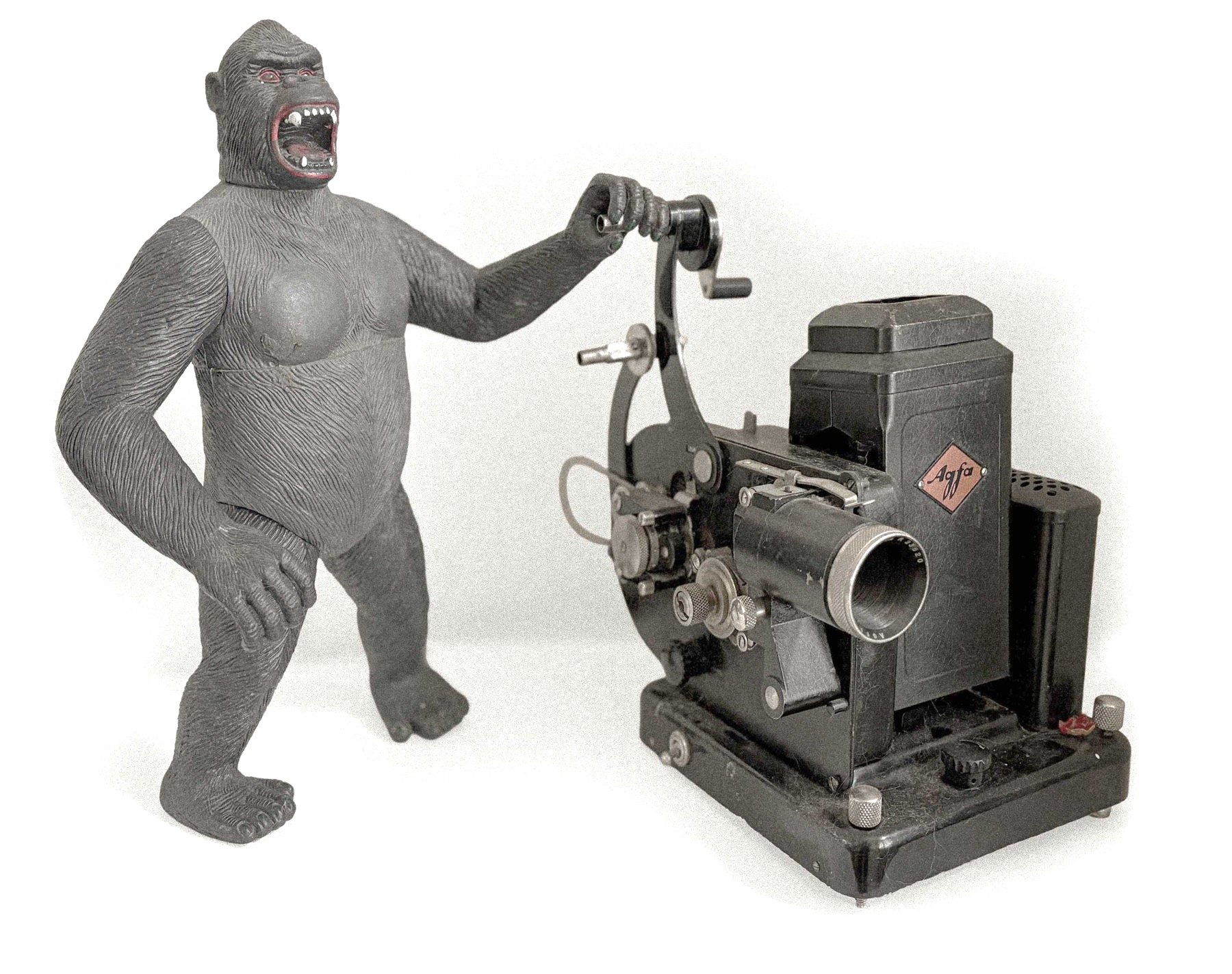 A vintage King Kong figure cranking the handle of an Agfa movie projector.