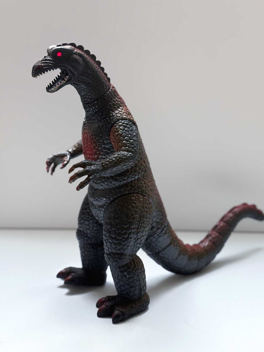 A Dor Mei vinyl kaiju figure in profile, showing a row of low rounded nubs along the neck and back instead of Godzilla's tall triangular dorsal plates, and one red painted eye.