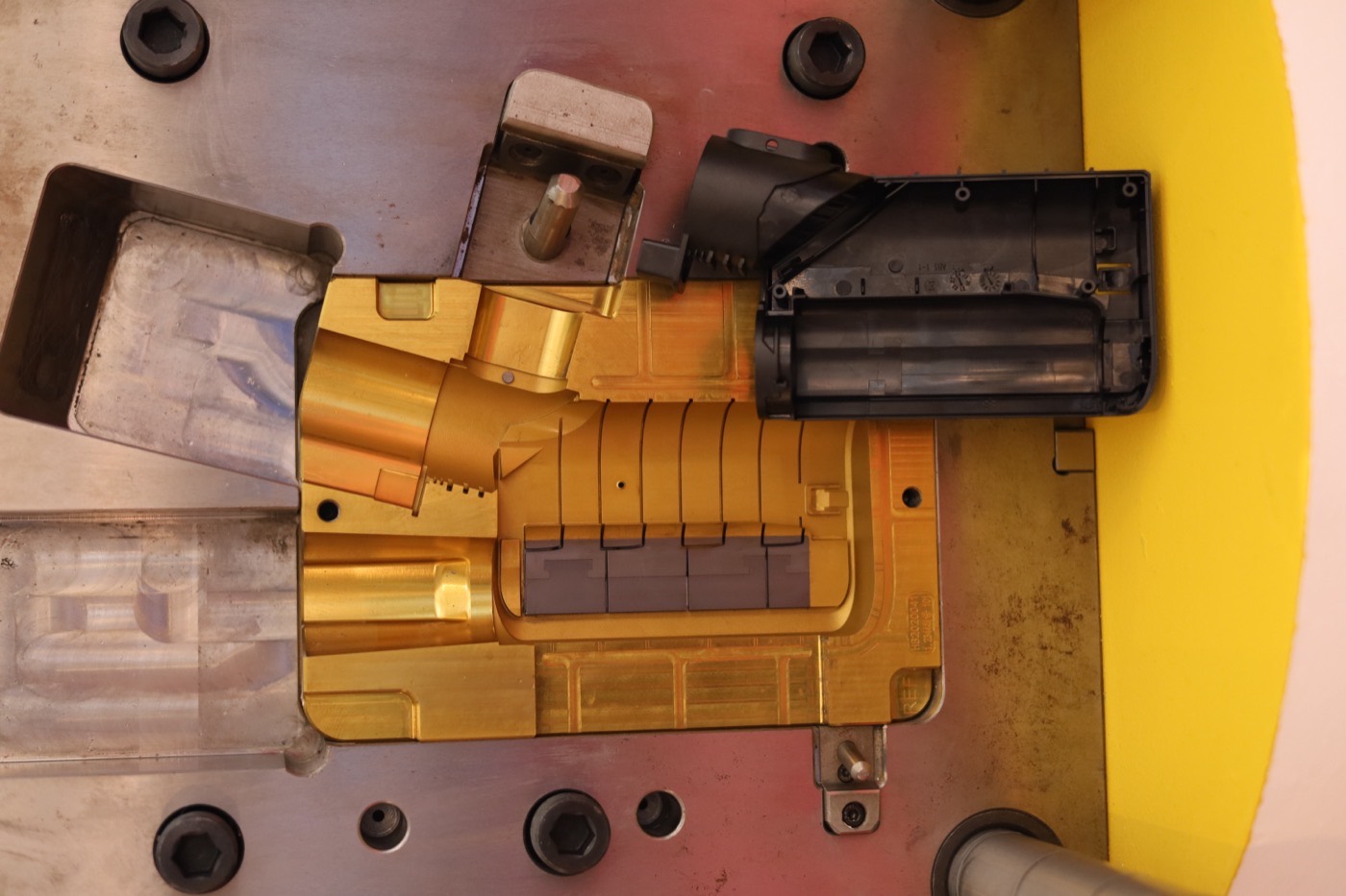One half of an open steel injection mould, the polished cavity and its slides, with the moulded plastic part lying beside it
