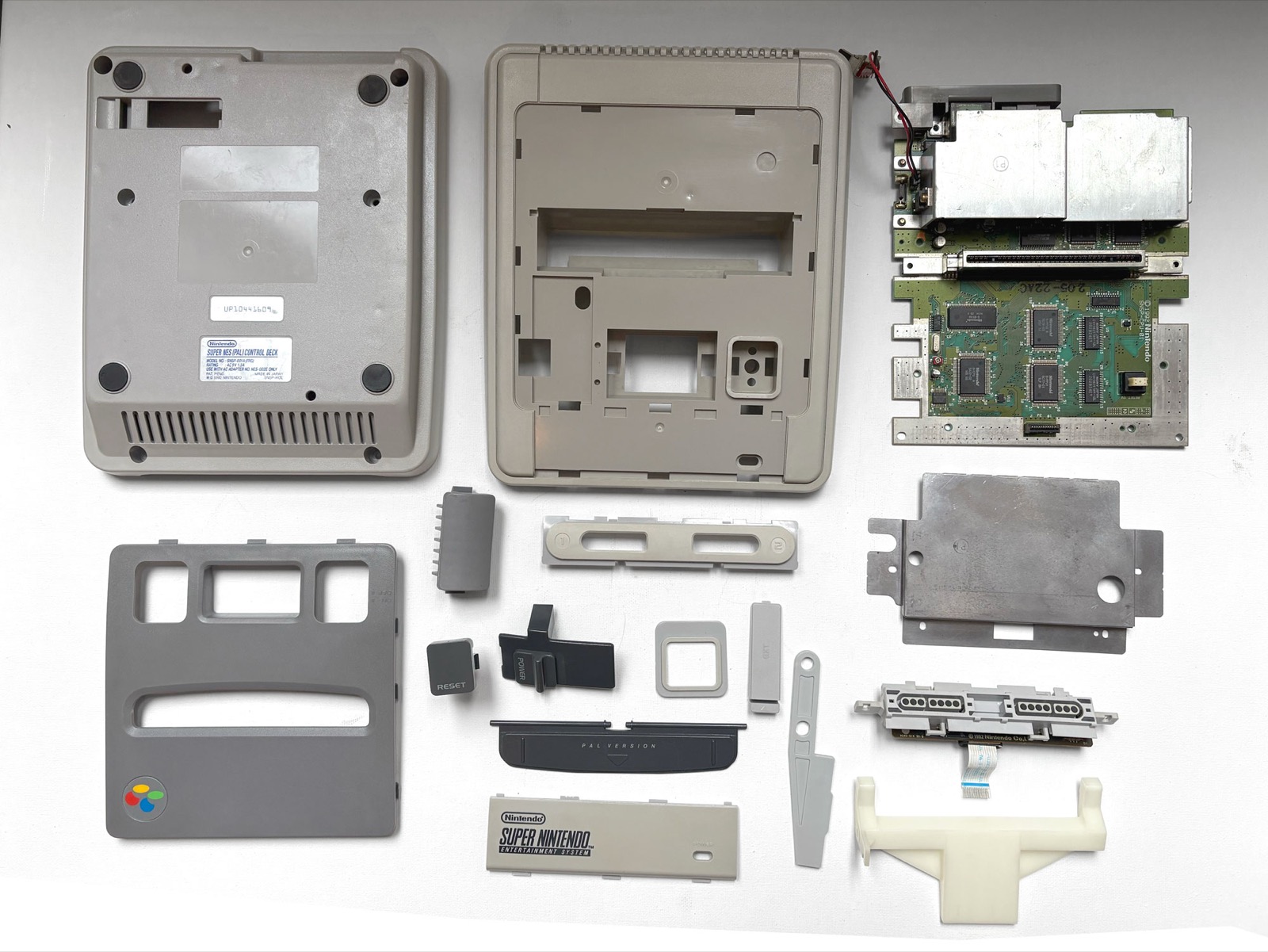 The same Super Nintendo parts laid out flat after treatment. The khaki has gone and the mouldings read as cool grey, with the two-tone grey of the original design visible again between the body and the top panel.