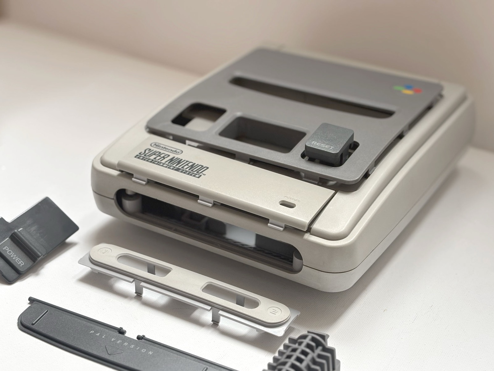 The console part-assembled after treatment, the grey body carrying the darker grey top panel, with the Super Nintendo badge plate and the PAL VERSION strip laid out in front of it.