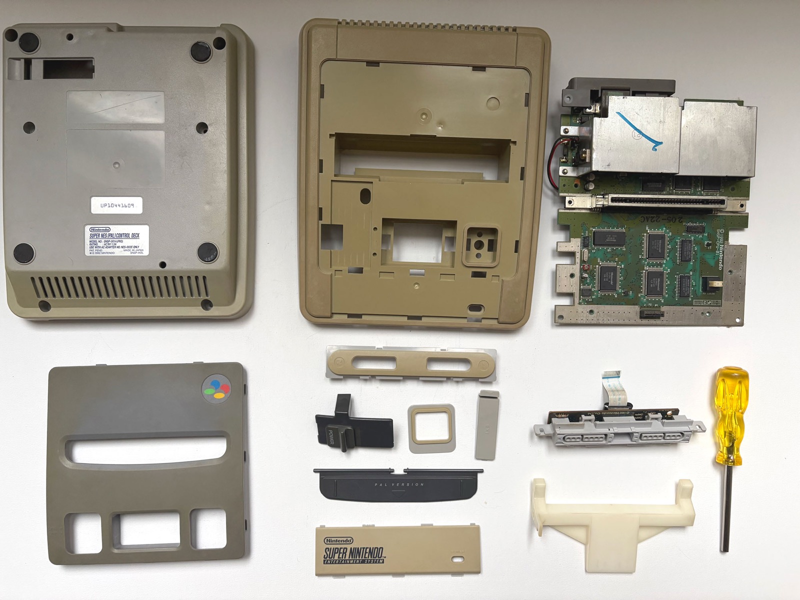A Super Nintendo PAL console stripped to its parts and laid out flat before treatment. Every moulded piece has gone to a deep khaki, darkest on the upper shell, while the internal white cartridge guide at the lower right is still almost white.