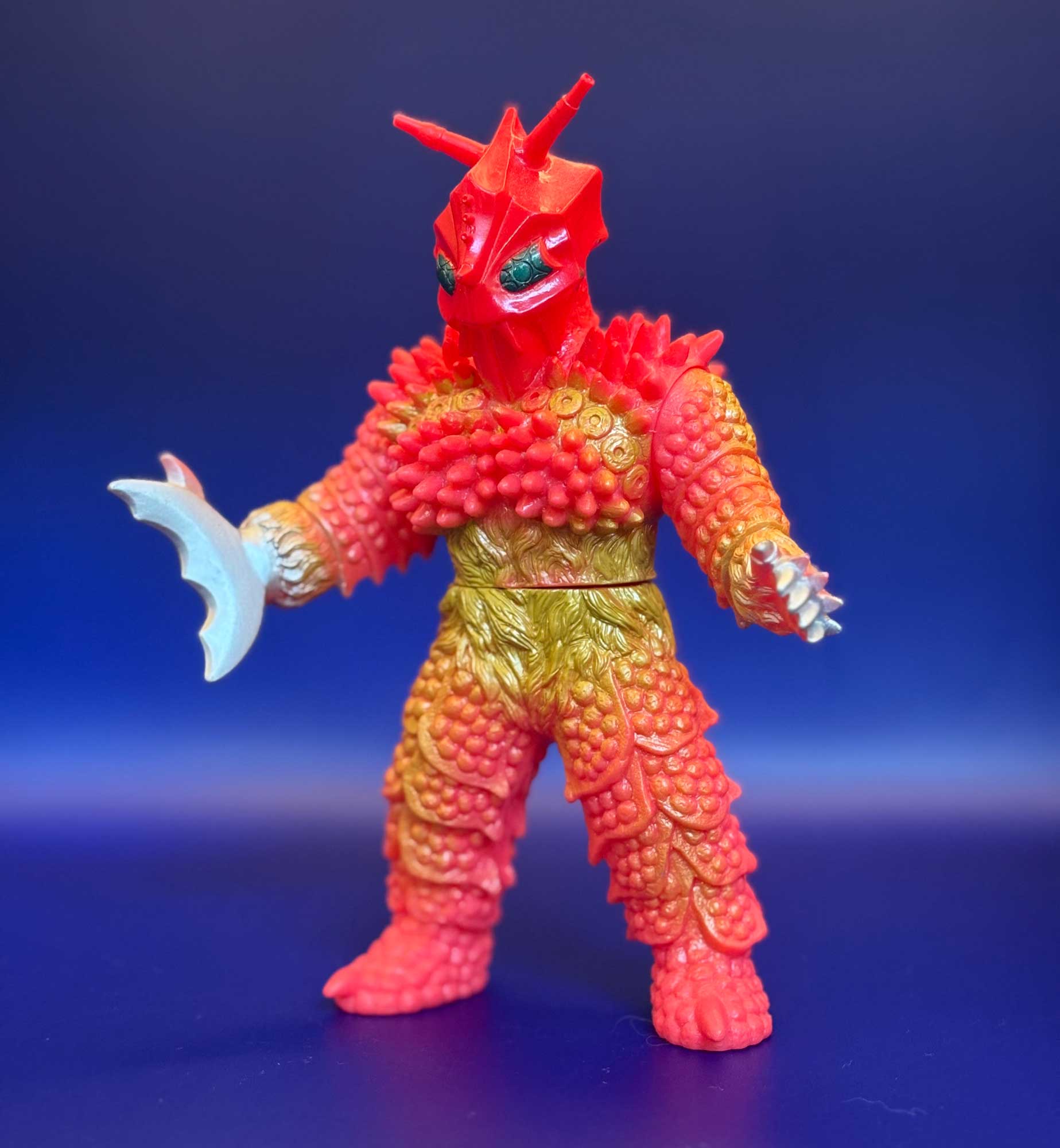 Una figura kaiju in vinile morbido vista di fronte su fondo blu: due lunghe antenne a corno, occhi verdi, un corpo di protuberanze rosa e arancio che sfumano nell'oliva alla vita, una lama pallida a mezzaluna su un avambraccio e una mano artigliata sull'altro
