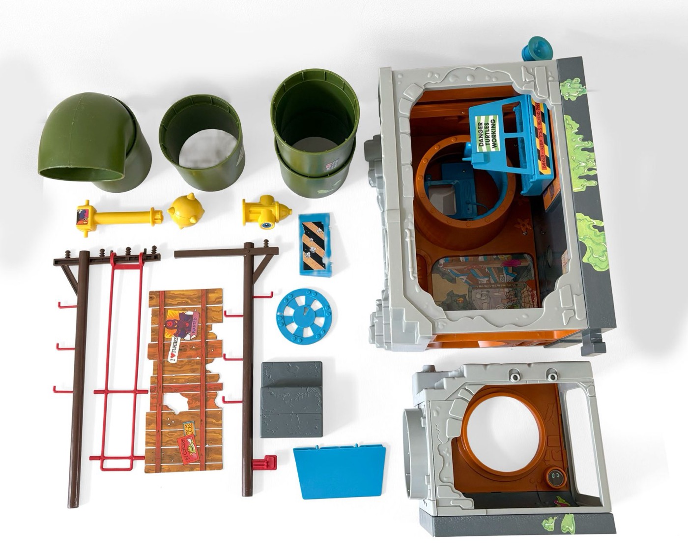 All parts of the Sewer Playset laid out flat: three green sewer pipe sections, two telephone poles with the red battle swing, the printed fence backdrop, barricade, manhole cover, fire hydrant, periscope and both building sections